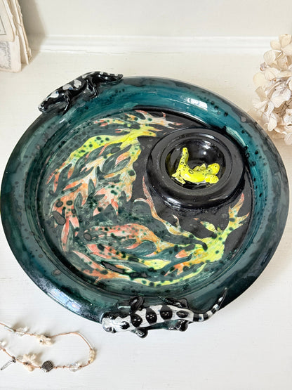 A decorative ceramic fish bowl with a glossy dark teal rim, hand-painted koi fish pattern in green, orange, and black, and a small black central pond section containing a bright yellow frog figurine. The bowl is styled on a white surface with dried flowers and a beaded necklace nearby.