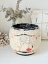 A vintage-style ceramic planter pot with raised figures and floral details, featuring a distressed cream finish with black and red accents, styled on a white surface with dried flowers and papers in the background.