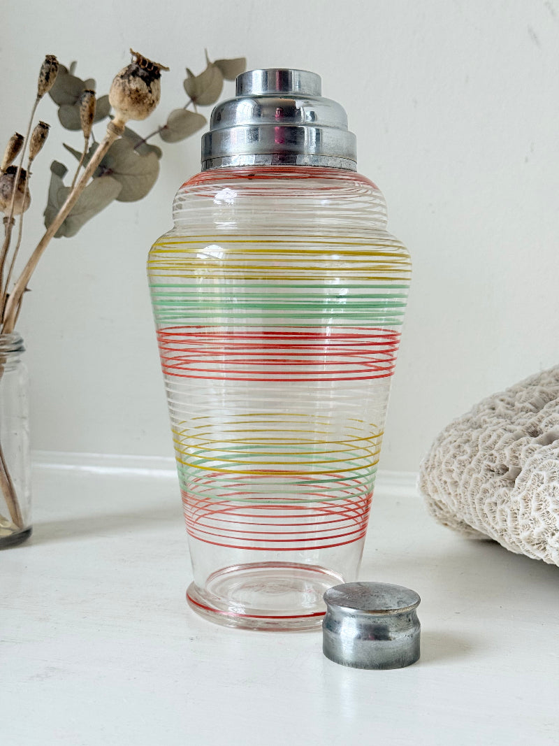 Vintage glass cocktail shaker with multicoloured stripes and silver lid photographed on a white surface.