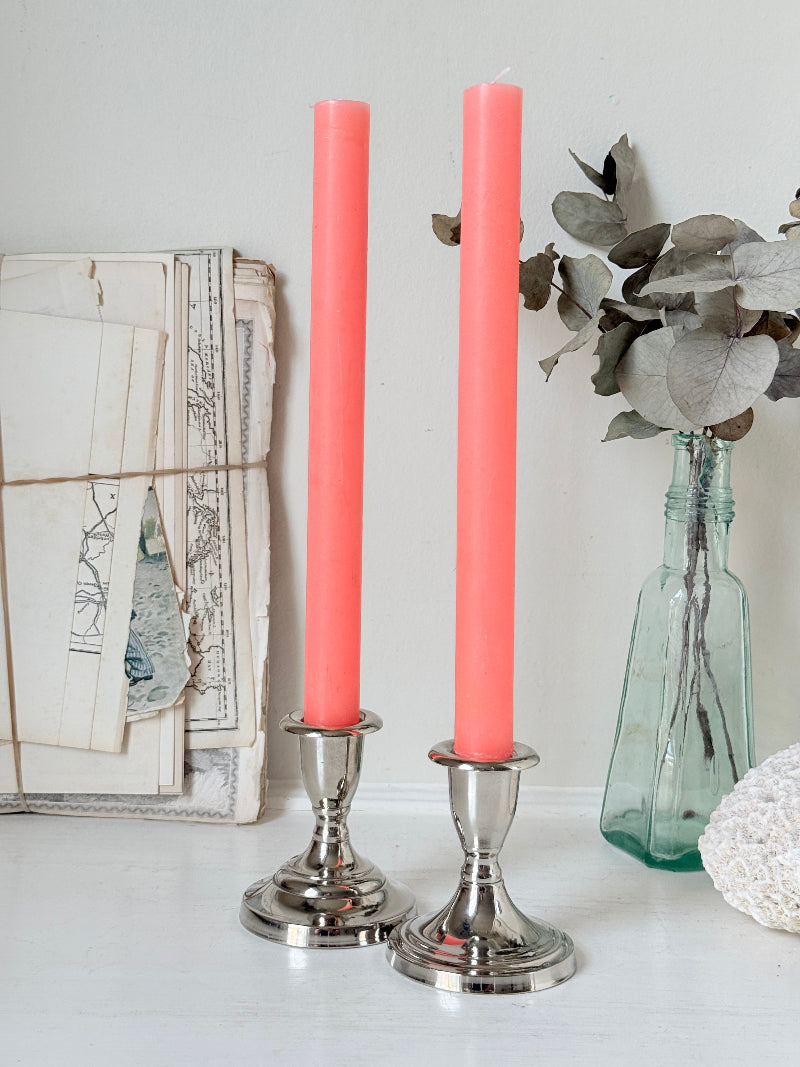 Pair of silver tone candlesticks with urn sconces and round stepped bases holding coral candles, photographed on a white surface