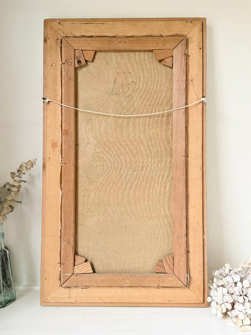 A rustic, vintage-style wooden picture frame with a linen backing board, photographed standing upright against a neutral off-white wall with decorative dried flowers on either side.