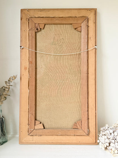 A rustic, vintage-style wooden picture frame with a linen backing board, photographed standing upright against a neutral off-white wall with decorative dried flowers on either side.