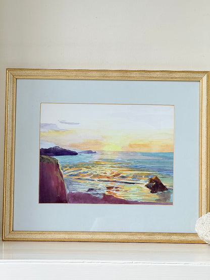 Framed coastal watercolour of sunset over sea with rocks in foreground, in gilt frame with pale mount, photographed on white surface.