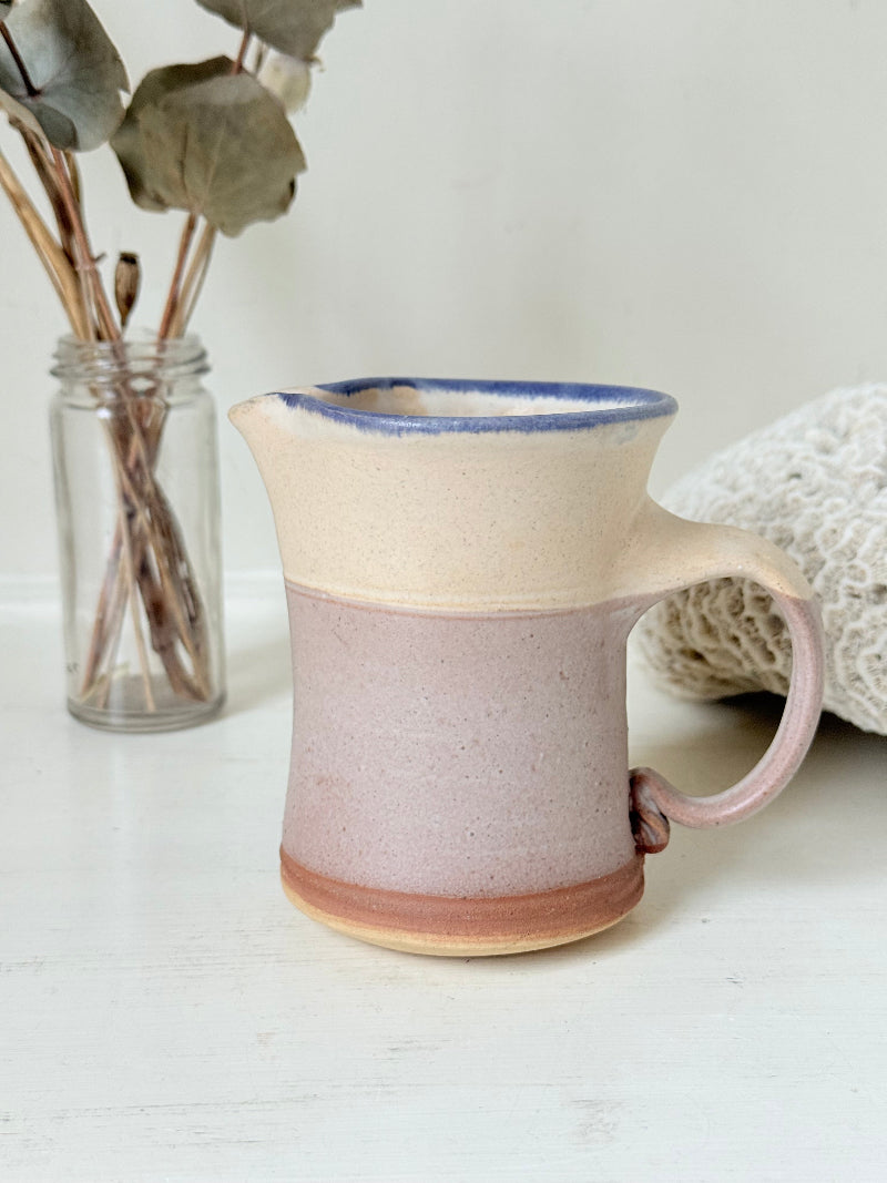 Studio pottery milk jug in cream and lavender glaze with impressed initials, photographed on a white surface.