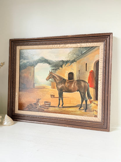 A framed vintage oil painting depicting a brown horse standing in a stable courtyard, set in a dark brown wooden frame on a white surface against a light beige wall.