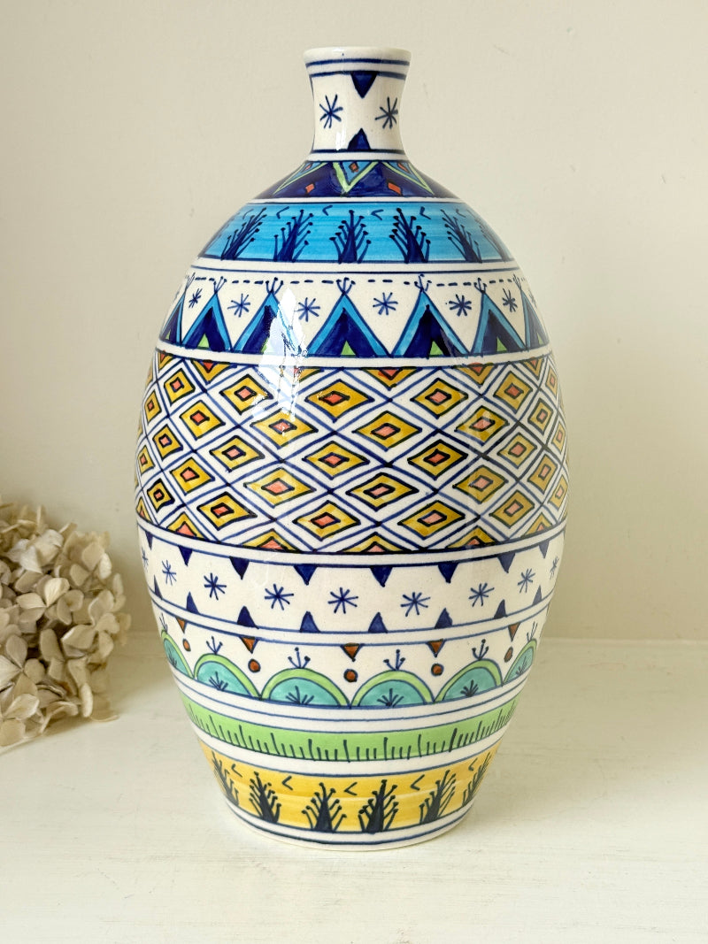 A hand-painted ceramic vase with a colorful Mediterranean-style geometric pattern, featuring blue, yellow, green, and white designs, set against a neutral cream background.