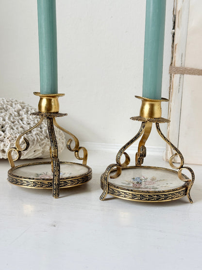 A pair of vintage-style gold and bronze candlestick holders with floral porcelain bases, shown on a white surface with mint green taper candles.