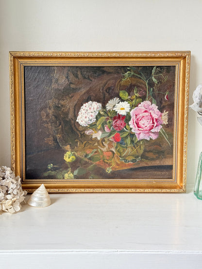A framed still life oil painting featuring a bouquet of pink, white, and yellow flowers, housed in an ornate gold frame, displayed on a white shelf with decorative seashells.