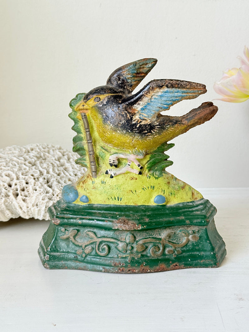 Vintage cast iron bird bank with a painted bird perched on a green decorative base, set against a light background with a lace doily and a flower prop.
