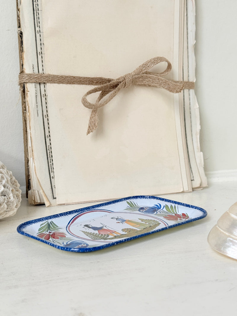 A small rectangular ceramic tray with a blue rim and a colorful illustrated scene on the front, styled on a white surface in front of stacked vintage books tied with twine.