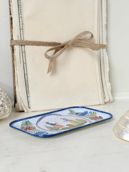 A small rectangular ceramic tray with a blue rim and a colorful illustrated scene on the front, styled on a white surface in front of stacked vintage books tied with twine.