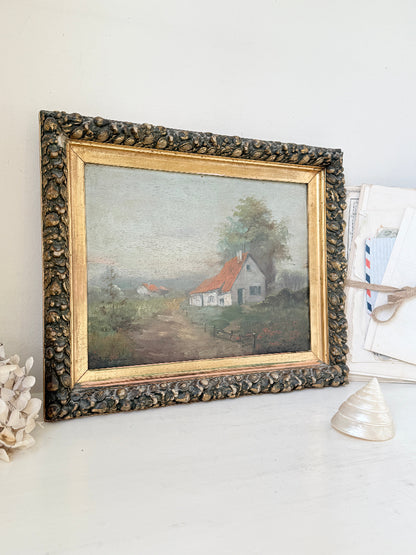 An antique framed landscape oil painting featuring a small white house with a red roof, set in a rustic dark wood frame with a gold inner border, displayed on a white surface.