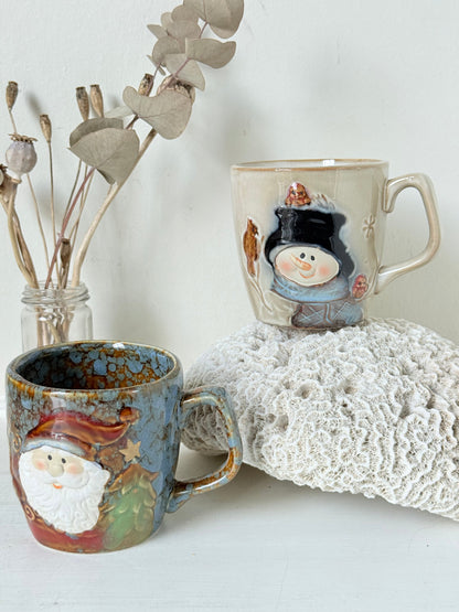 Pair of Christmas mugs with Santa and snowman designs photographed on a white surface.