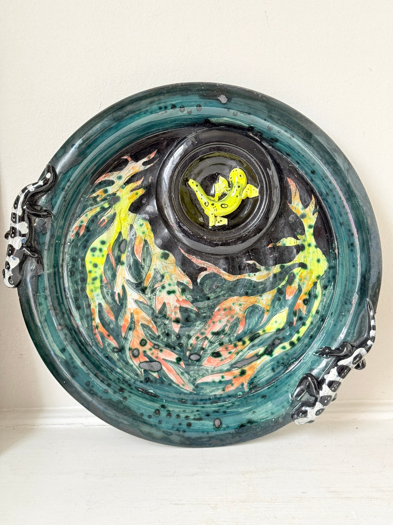 A round ceramic plate with a glossy teal and black glaze, decorated with colorful abstract fish designs in yellow, orange, and green, and featuring black-and-white spotted handles on the sides.