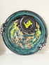 A round ceramic plate with a glossy teal and black glaze, decorated with colorful abstract fish designs in yellow, orange, and green, and featuring black-and-white spotted handles on the sides.