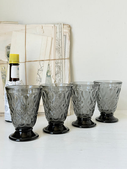 Set of four smoke grey pressed glass footed goblets with a diamond pattern, photographed on a white surface