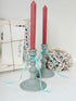 Two distressed sage green candlestick holders with red taper candles, decorated with light blue curly ribbon, styled with vintage postcards and a dried flower on a white surface.