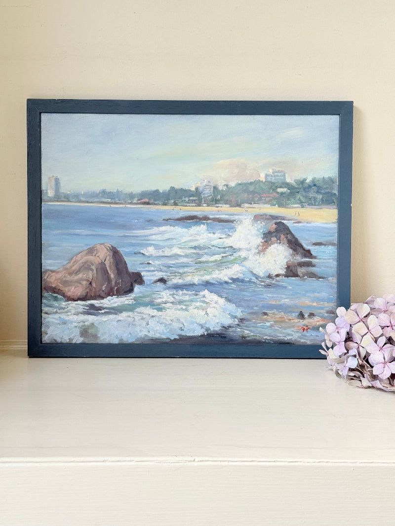 Original oil painting of waves crashing against rocks, signed seascape in slate blue wooden frame on white surface

