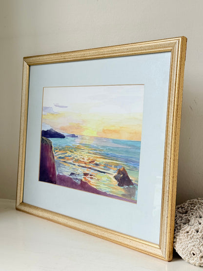 Framed coastal watercolour of sunset over sea with rocks in foreground, in gilt frame with pale mount, photographed on white surface.