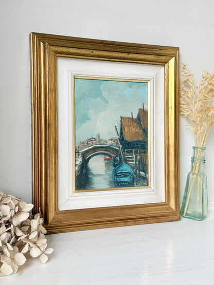 A framed art print depicting a Venetian canal scene with a gondola and bridge, styled on a white surface with dried flowers and a glass vase.