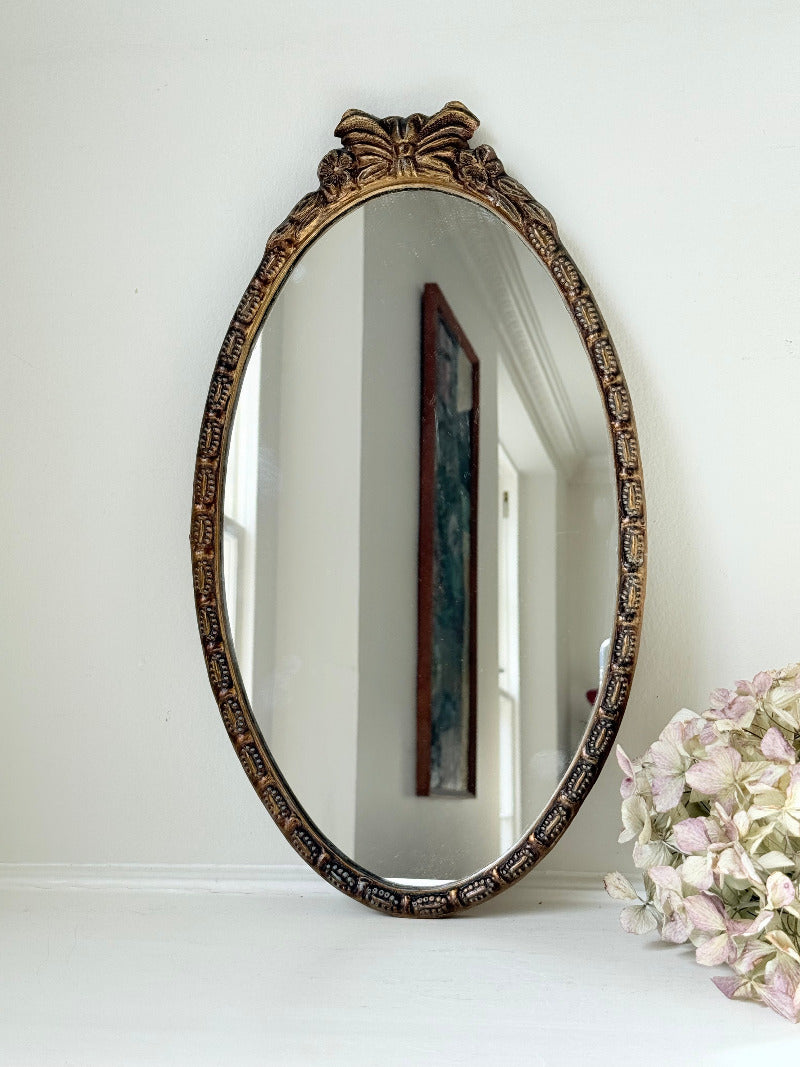 Vintage oval wall mirror with decorative gilt frame and bow crest, styled against a neutral wall with hydrangeas.