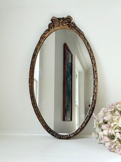 Vintage oval wall mirror with decorative gilt frame and bow crest, styled against a neutral wall with hydrangeas.