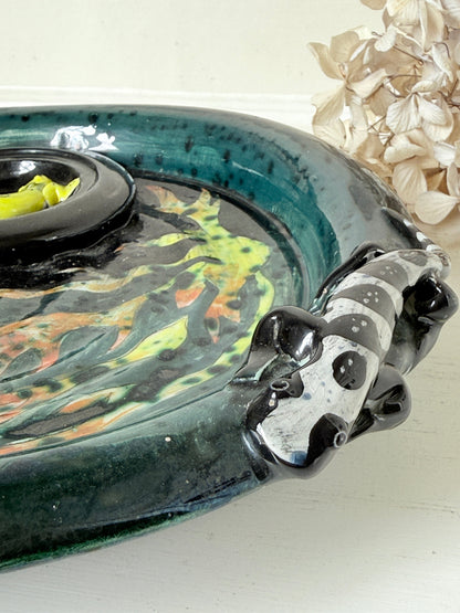A close-up of a decorative ceramic tray with a glossy teal and black glaze, featuring a black circular insert and a sculpted black-and-white handle shaped like a stylized animal head, set against a light background with dried flowers.