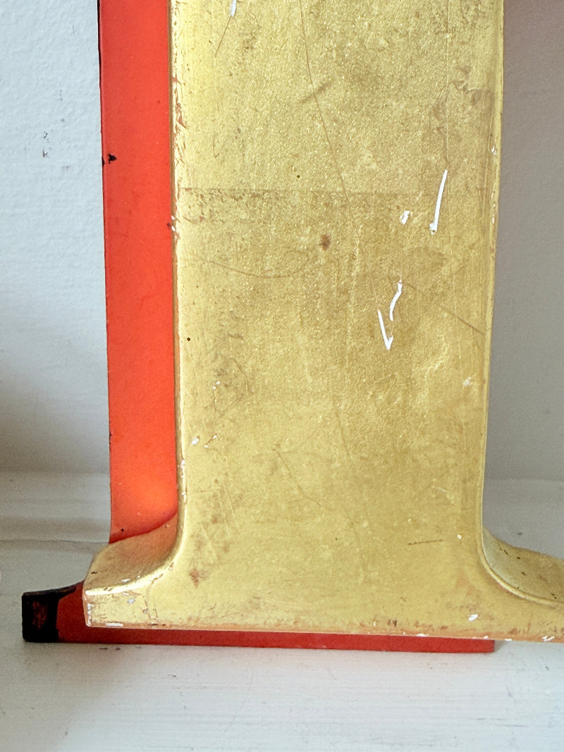 A close-up of a vintage wooden object, likely a record player turntable or speaker cabinet, showing a distressed gold-finished top surface and a bright red-orange side panel.