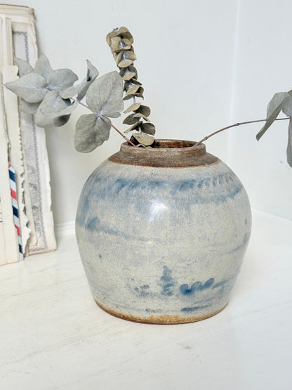 A round handmade ceramic vase with a blue and white speckled glaze and a rustic brown rim, holding dried eucalyptus stems, set on a white surface against a white background.