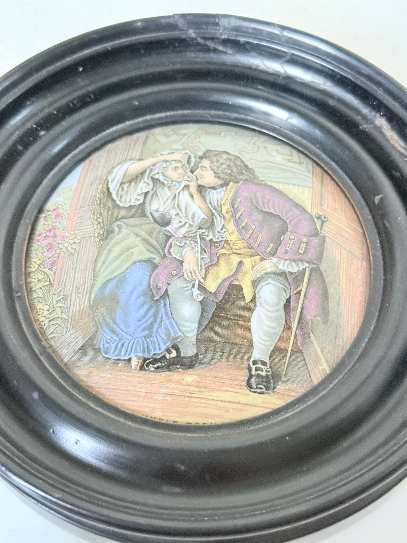 Antique Prattware pot lid in a black round frame with a detailed Victorian scene on a white surface.