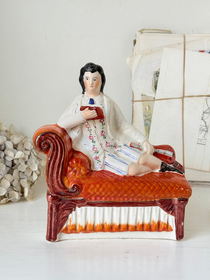 A vintage ceramic figurine of a man reclining on an orange-red chaise lounge, set against a neutral background with decorative dried flowers and papers.