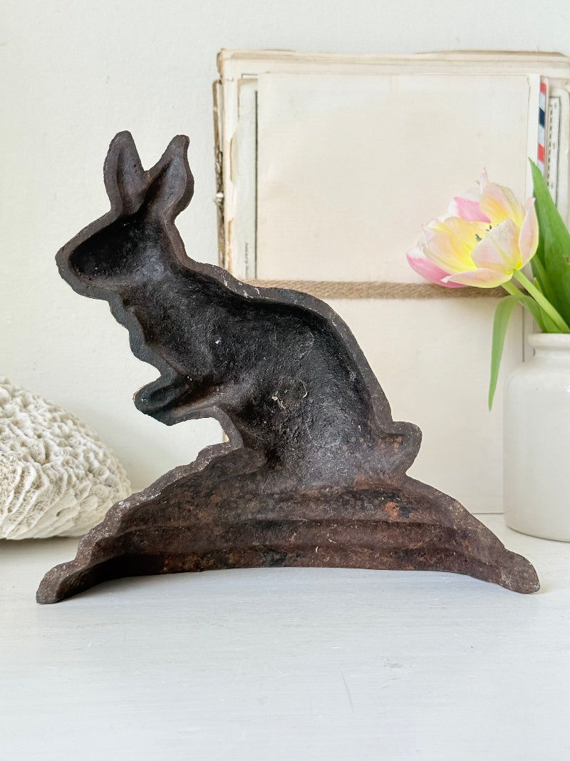 A rustic cast iron bunny-shaped dish or planter with a dark textured finish, displayed on a white surface with a white vase of tulips and a neutral background.