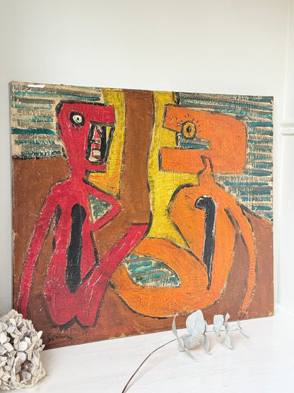 An abstract canvas painting displayed on a white surface against a white wall, featuring bold brushstrokes in red, orange, yellow, brown, and teal. Decorative white dried flowers and a textured white object sit at the base of the canvas.