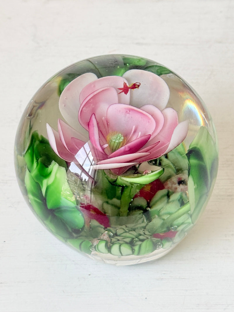 Large clear glass paperweight with a pink flower and green leaves inside, photographed on a white surface