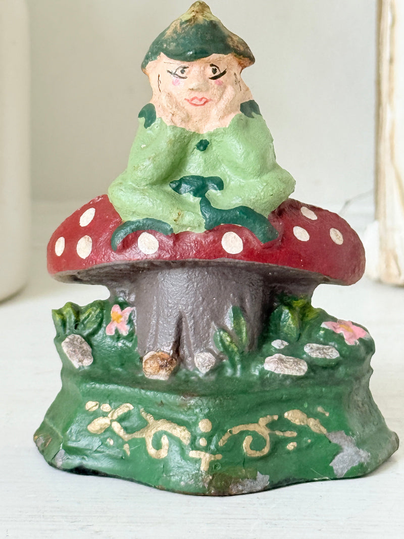 A vintage hand-painted cast iron gnome figurine sitting on a red and white spotted mushroom, set against a light-colored background.