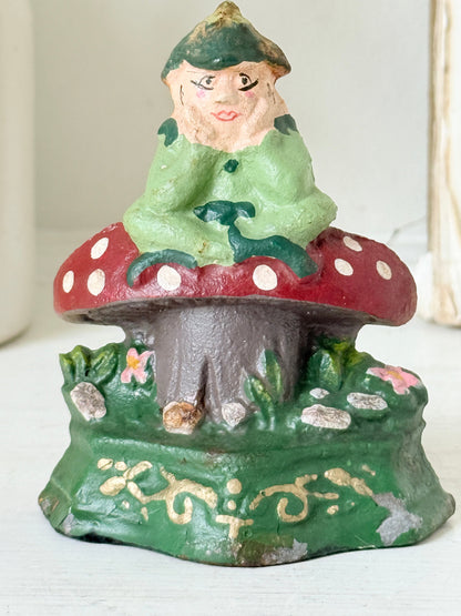 A vintage hand-painted cast iron gnome figurine sitting on a red and white spotted mushroom, set against a light-colored background.