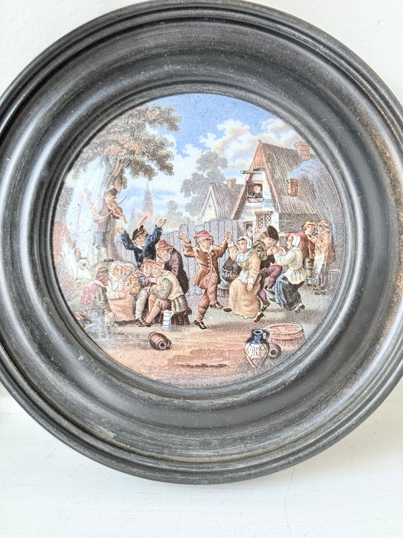 Antique Prattware pot lid in black round frame with rural village scene on white surface.