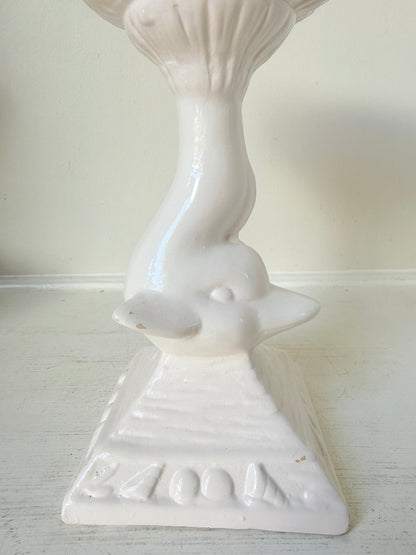 A white ceramic bird-shaped candlestick holder with a pedestal base, photographed against a plain off-white wall and surface.