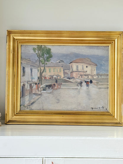 A framed impressionist landscape painting depicting a street scene with buildings, a tree, and figures, housed in a wide gold frame and displayed on a white surface against a light wall.