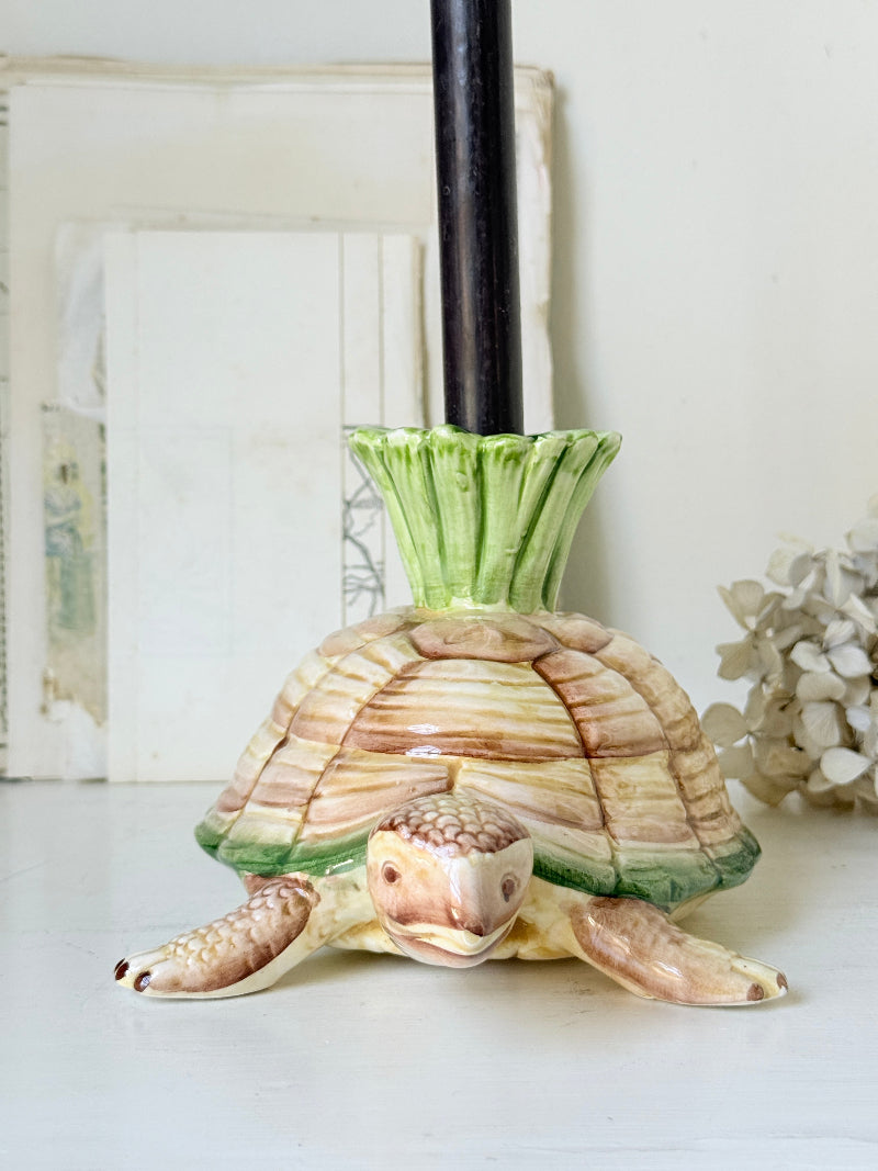 A ceramic turtle-shaped candle holder with a green leafy top and a black candle inserted, set against a soft, off-white background with decorative elements.