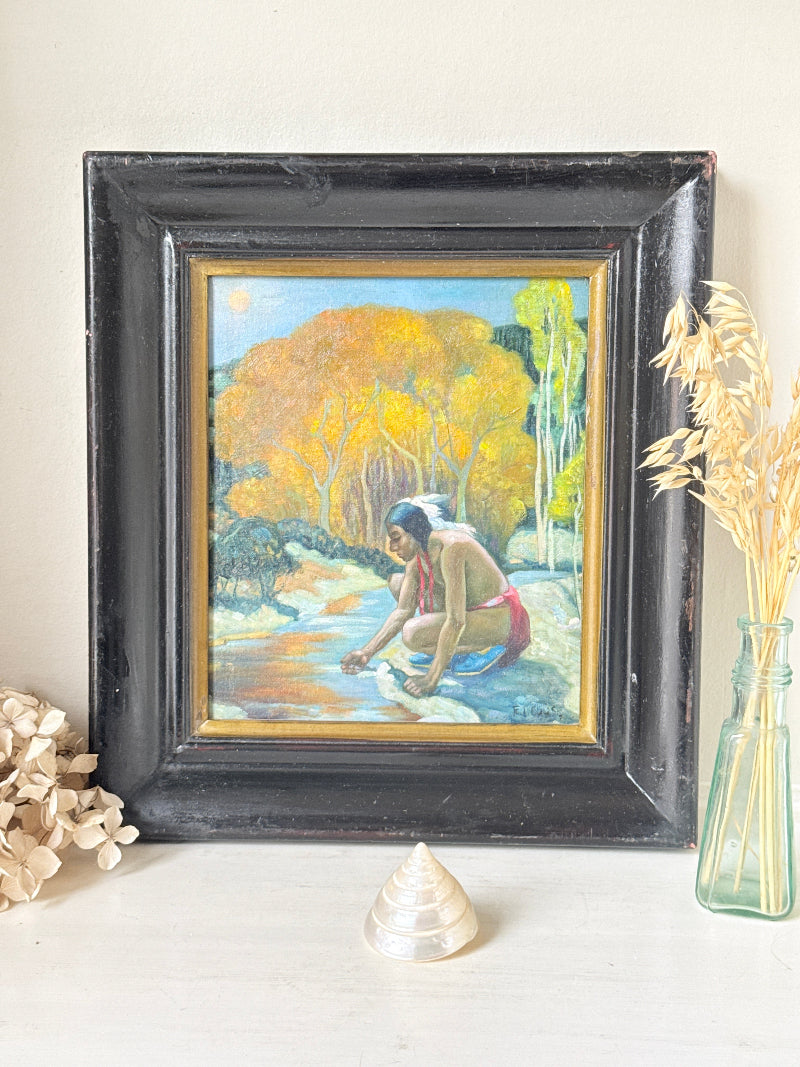 A framed painting depicting a Native American figure by a river, styled on a tabletop with dried flowers and a glass vase against a neutral wall.