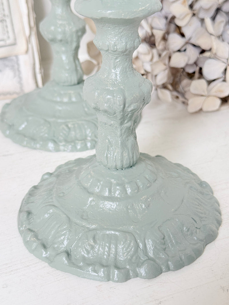 A close-up of a distressed, light sage green cast iron candlestick base with ornate embossed detailing, set against a softly blurred background of another matching candlestick and dried flowers.