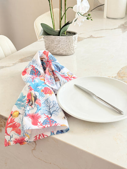 Coral reef print cotton tea towel in crisp white with colourful coral and tropical fish motifs, photographed indoors with a white plate