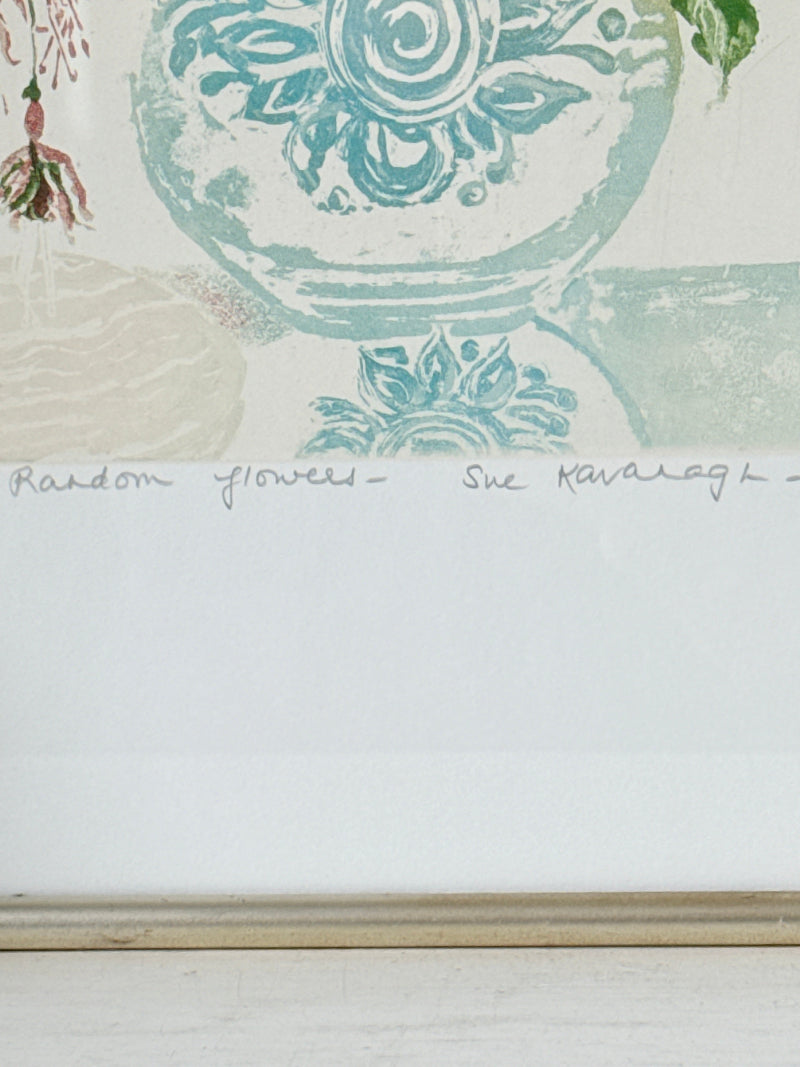 A close-up of a framed art print featuring a light blue and green floral design on a white background, with a handwritten signature and the text &