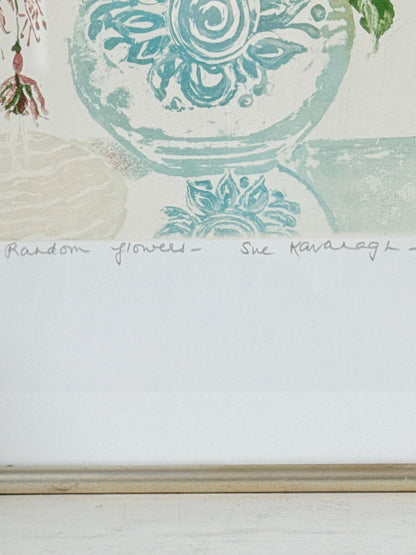 A close-up of a framed art print featuring a light blue and green floral design on a white background, with a handwritten signature and the text &