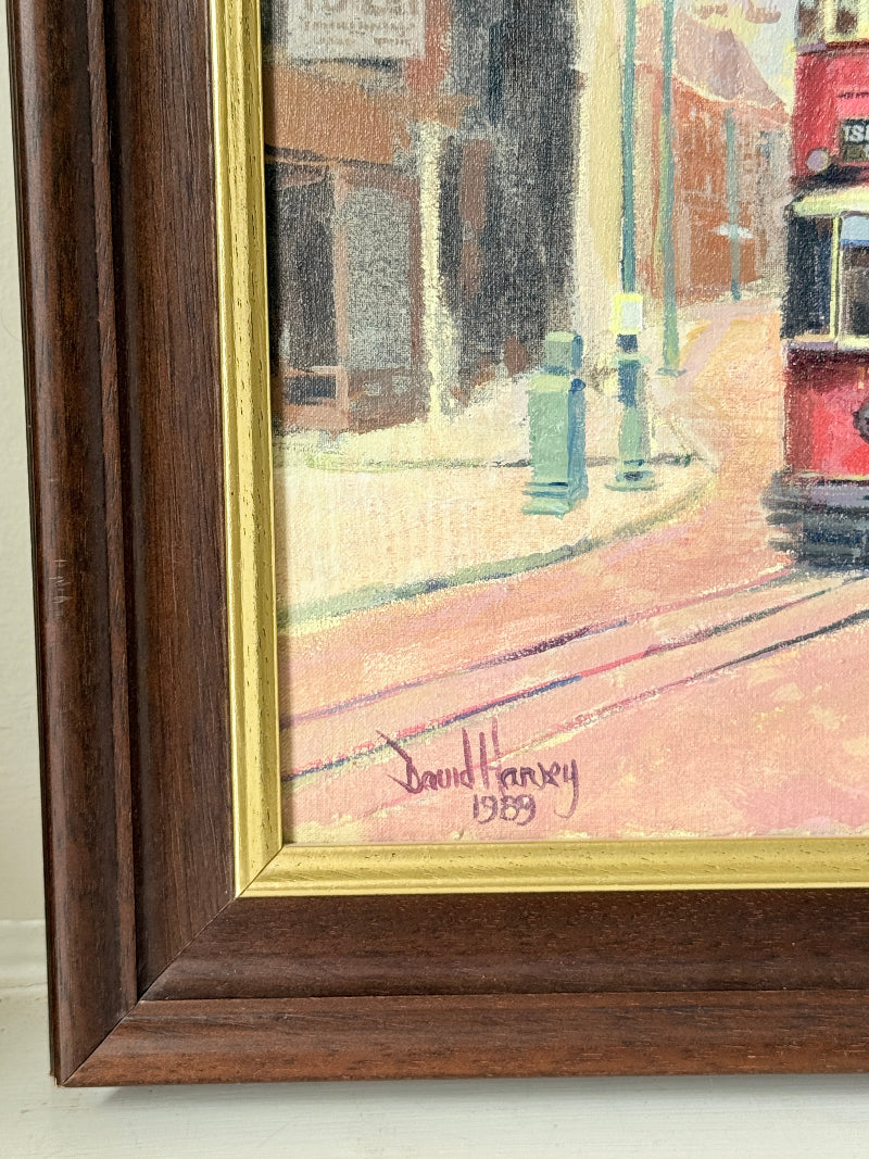 A framed painting showing a street scene with a red tram, painted in warm tones, with a gold inner frame and a dark brown wooden frame. The artist&