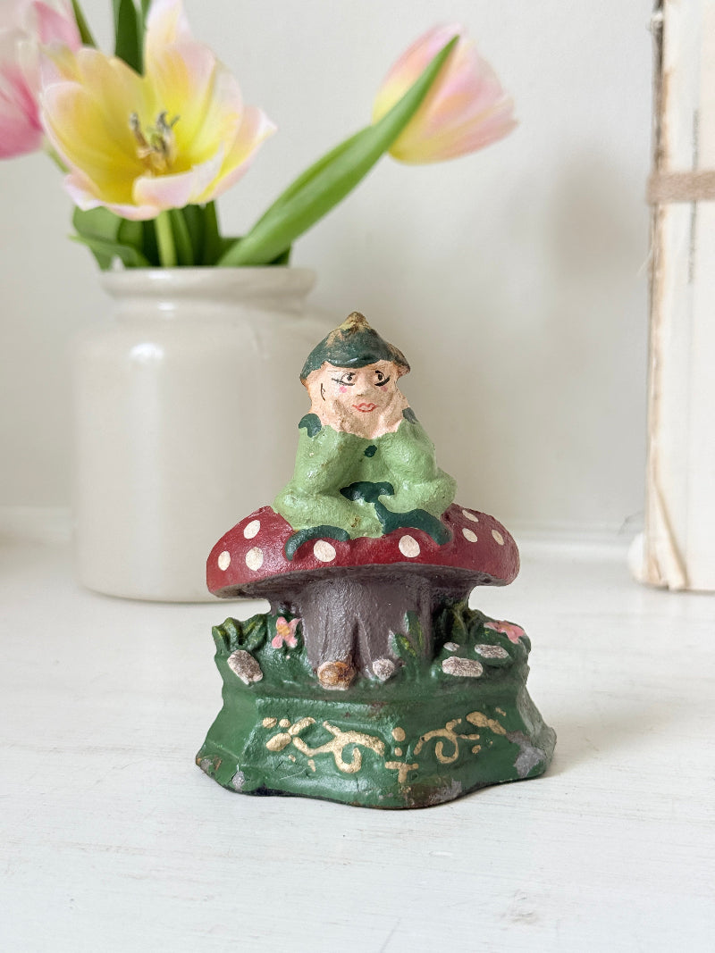 A small decorative gnome figurine sitting on a red-and-white spotted mushroom cap, placed on a white surface with a blurred white vase of pink tulips in the background.