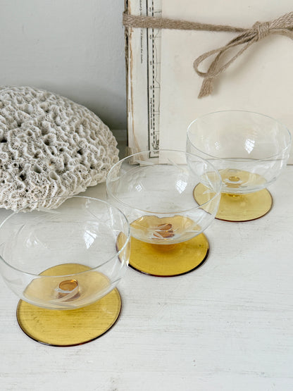 Three clear glass coupe-style glasses with round amber-yellow bases, arranged on a white tabletop with a crocheted textile and a rustic box in the background.