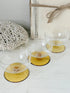 Three clear glass coupe-style glasses with round amber-yellow bases, arranged on a white tabletop with a crocheted textile and a rustic box in the background.