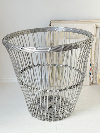 A large, round, open-top metal wire basket with a woven basket-weave pattern and a solid metal base, shown on a white floor against a white wall with some vintage picture frames in the background.